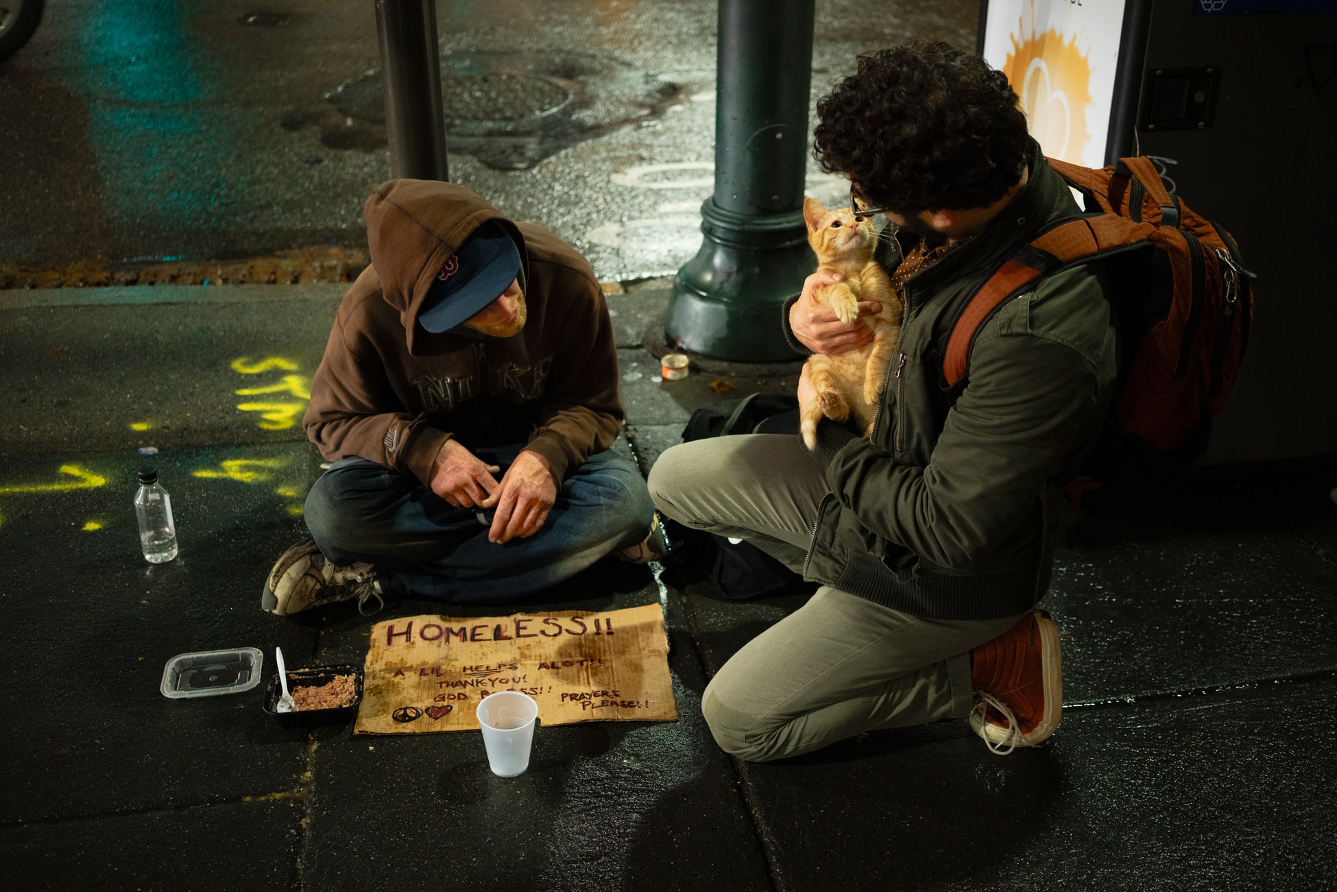 A photo of a homeless man sitting on the sidewalk with a sign saying "Homeless! A Little Helps a lot. Thank you, God bless. Prayers please!" A pedestrian is kneeling beside him petting his kitten and having a conversation with the homeless man.